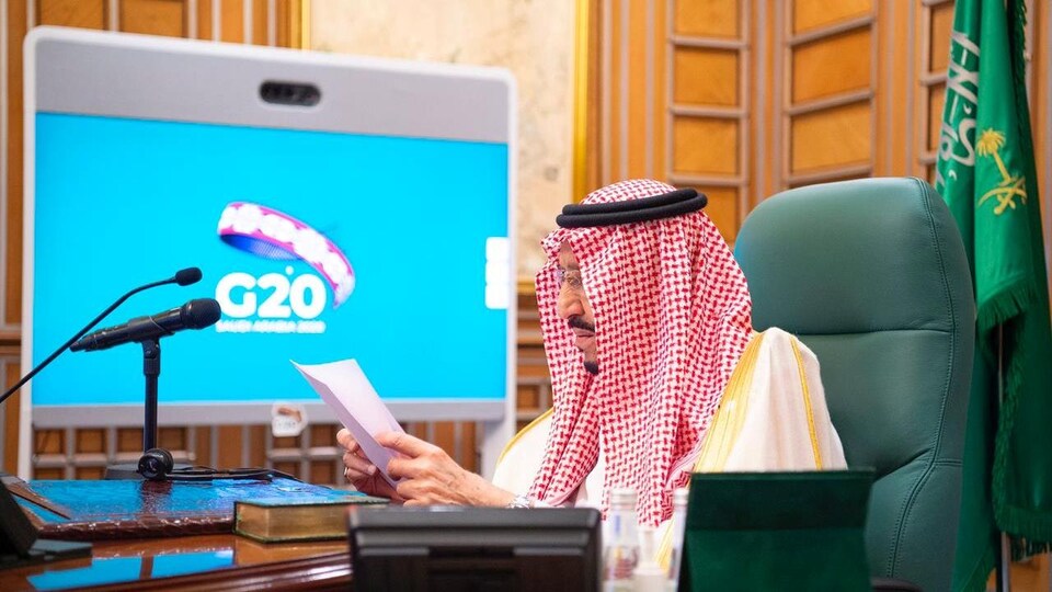Salmane ben Abdelaziz Al Saoud regarde un document. Un écran derrière lui affiche le logo du G20.