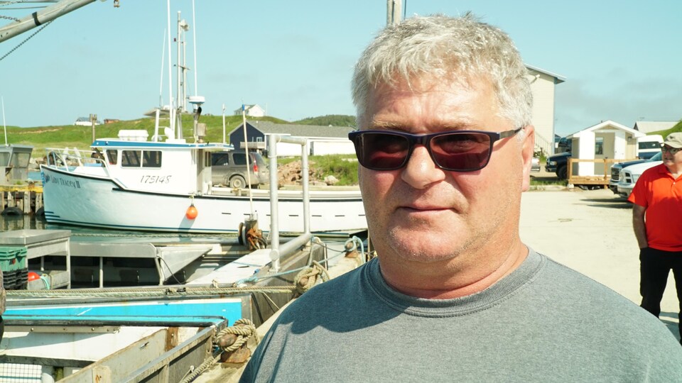 Un homme porte des lunettes fumées. Il est sur un quai dans un port. Un bateau de pêche au homard se trouve derrière lui. 
