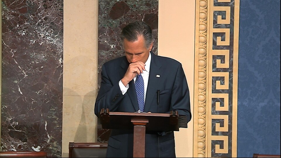 Mitt Romney, devant un lutrin, au Sénat, la main sur le bas du visage.