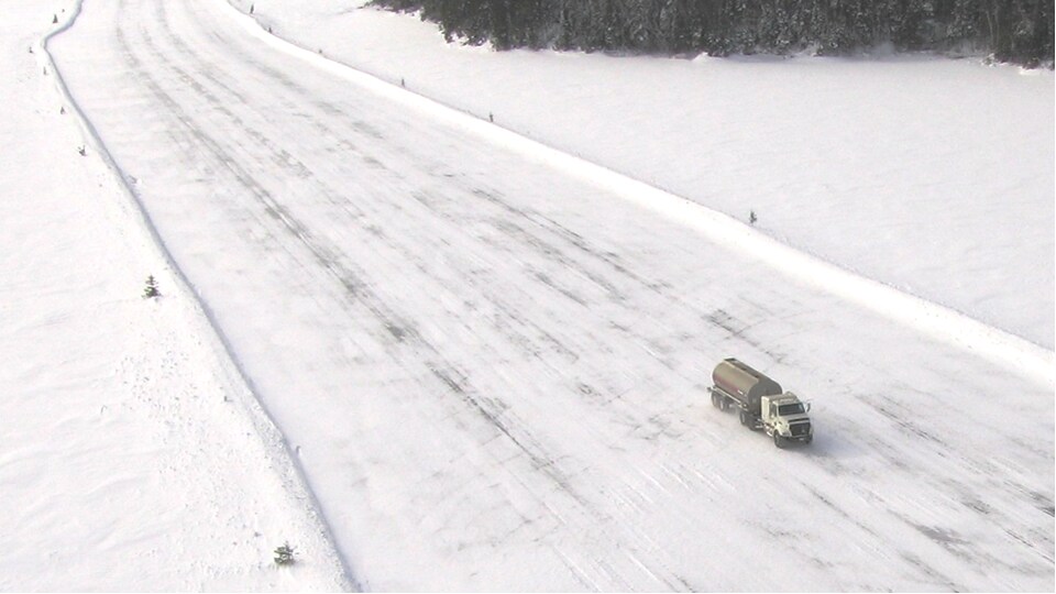 Un camion-citerne circule sur une route de neige et de glace au milieu au milieu d'une grande étendue de neige et de sapins.