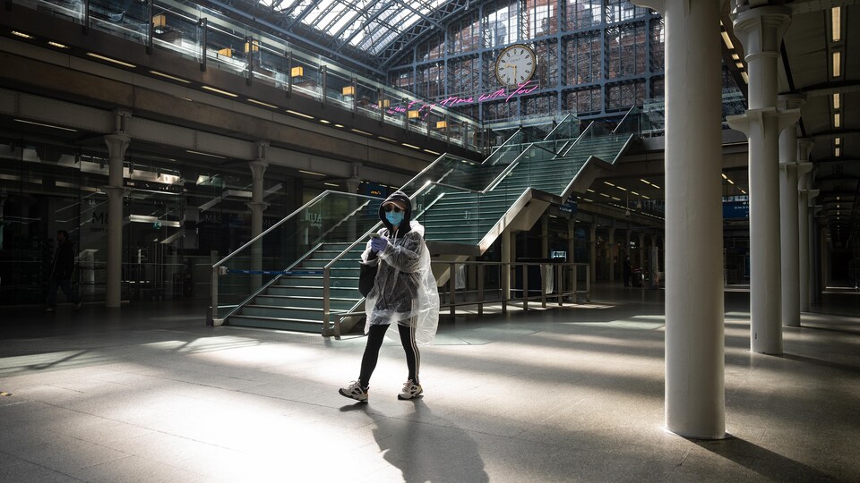 Une femme portant un masque, un poncho, une casquette et des verres fumés marche, seule, dans une gare.