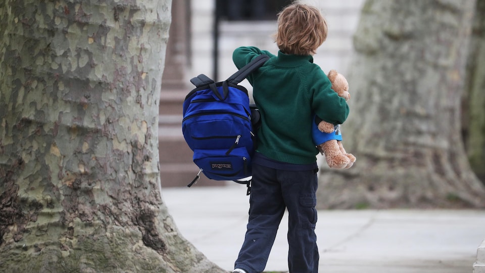 Un jeune garçon marche avec son sac d'école en main et sa peluche dans l'autre.
