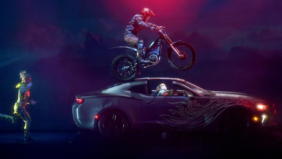 Un homme conduit une moto sur le toit d'une voiture qui se trouve sur scène.