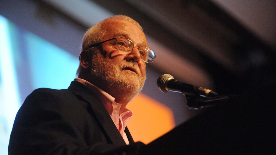 Russell Banks au micro, dans une salle sombre. L'homme a les cheveux blancs et porte des lunettes.