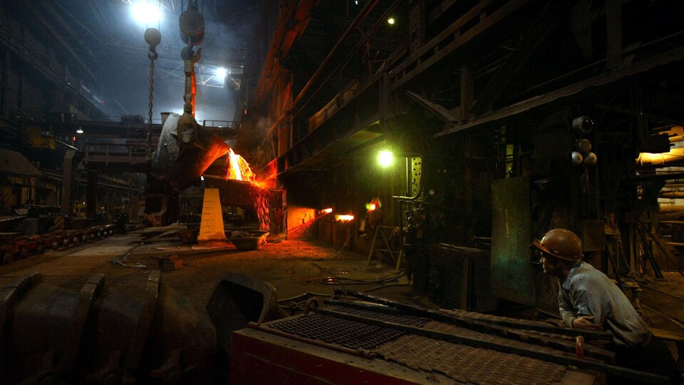 Un ouvrier avec un casque jaune observe de loin la fonte de métaux dans une usine.