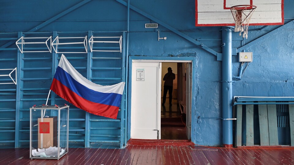 Un bureau de vote installé dans un gymnase. À l'avant-plan, un drapeau de la Russie et un panier de basketball.