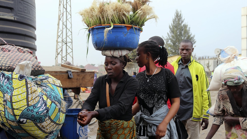 Une femme qui porte un panier rempli d'oignons verts sur sa tête se lave les mains. Des gens attendent en file derrière elle.