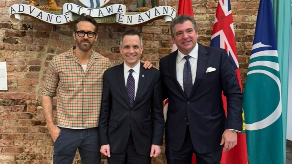 Ryan Reynolds pose devant les armoiries de la Ville d'Ottawa, une main autour de l'épaule du maire Mark Sutcliffe.