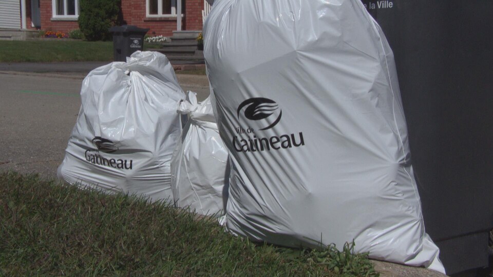 Des sacs de poubelle blancs avec le logo de la Ville de Gatineau, placés sur le bord d'une rue.