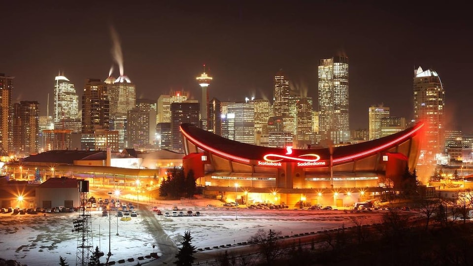 Une photo prise de nuit du Saddledome. En arrière-plan, des gratte-ciel sont illuminés.