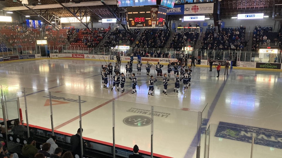 Des joueurs de hockey en cercle au centre de la patinoire lèvent leur bâton dans un aréna avec des spectateurs.