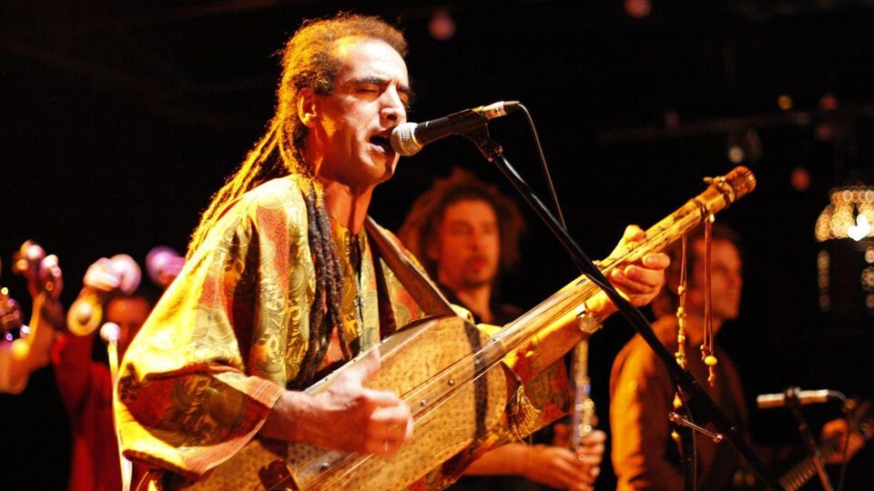 Un homme joue d'un instrument à corde en chantant dans un micro, avec quelques musiciens en arrière-plan. 