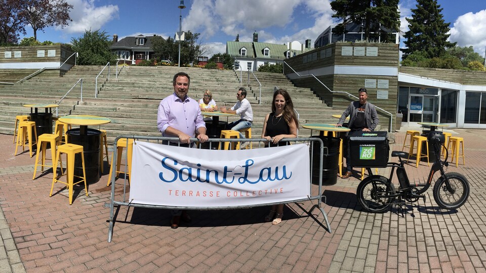 Le maire et la directrice se tiennent en arrière d'une affiche où il est écrit : Saint-Lau terrasse collective. Des tables les entourent. Ils sont dans le parc portuaire. En arrière plan des marches en bois.
