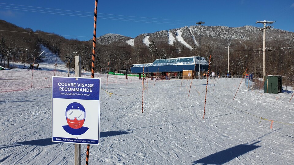 Vue d'ensemble d'une piste de ski. Au premier plan a été planté dans la neige un panneau d'information recommandant de porter un couvre-visage.