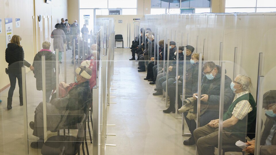 Des dizaines de personnes sont assises dans une salle d'attente, séparées par des plexiglas.