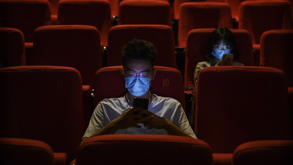 A man and woman wearing surgical masks are sitting in red chairs in a movie theater, staring at their smart phones. 