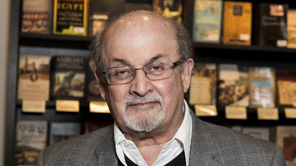 Lunettes sur les yeux et cheveux grisonnants, Salman Rushdie pose devant un mur couvert de livres.