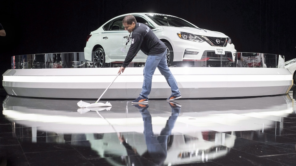 Un travailleur nettoie le sol devant un véhicule exposé.