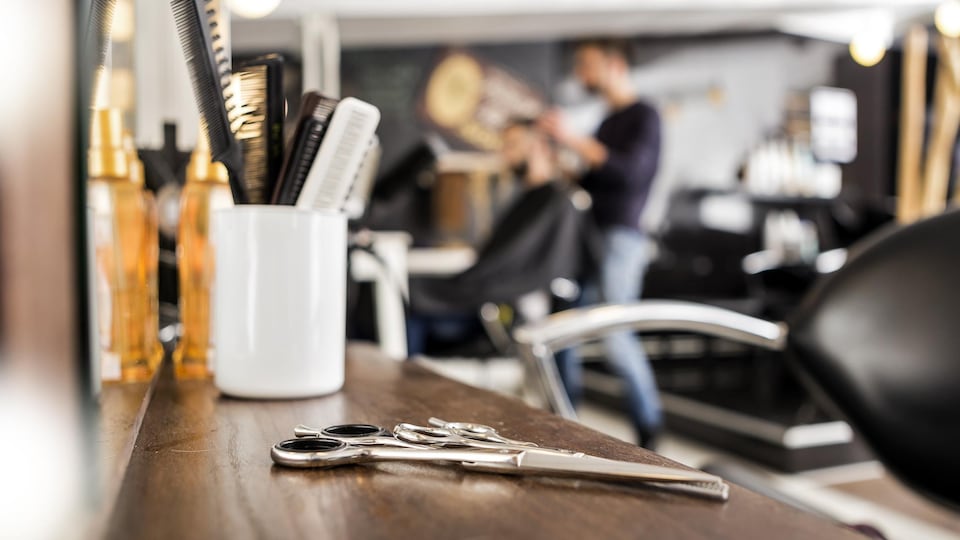 Gros plan sur des ciseaux de coiffure déposés sur un comptoir. À l'arrière-plan, un homme se fait coiffer par un barbier. 