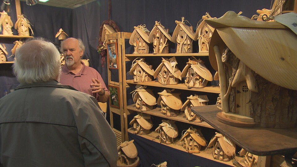 Un artisan du Salon des métiers d'art de Sherbrooke en discussion avec un visiteur. L'artisan fabrique de petites cabanes en bois très détaillées.