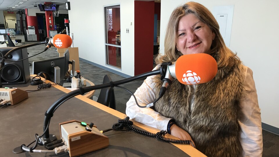 Portrait d'une femme dans un studio de radio. Elle est debout derrière une table sur lequel sont installés plusieurs micros.