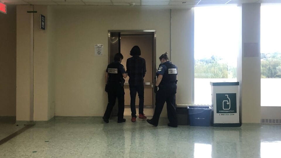 Samuel Cyr, de dos, entre dans un ascenseur. Il est menotté et est escorté par deux policiers.