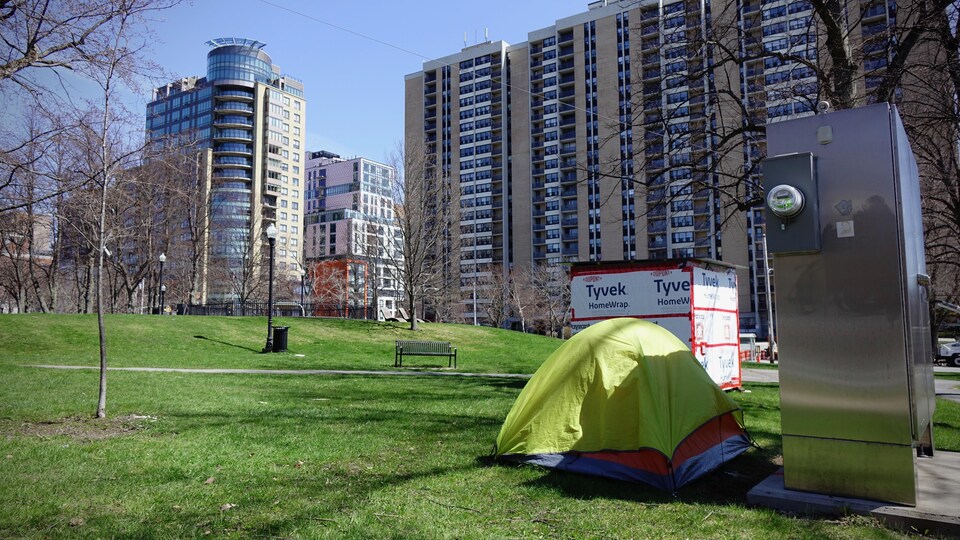 Un abri en bois et une tente dans un parc municipal, devant de hauts immeubles d'appartements.  