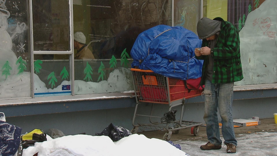 Un itinérant est devant un centre d'hébergement. Il est penché et tient un papier dans ses mains. Il est entouré de neige et d'un panier d'épicerie rempli couvert d'une toile bleue pour protéger ses effets personnels. 