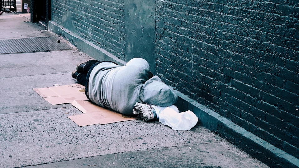 Un homme est allongé sur le trottoir, sur un morceau de carton le long d'un mur.