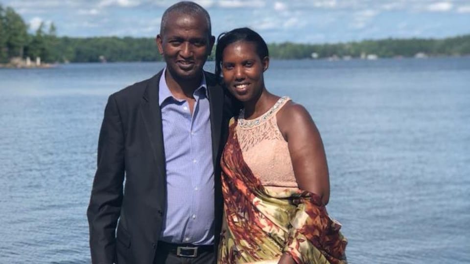 Un homme et une femme souriants pose sur un ponton sur un lac. Il porte un costume, elle un vêtement traditionnel africain