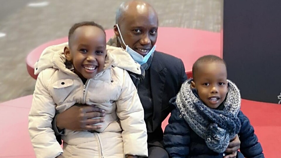 Un homme pose avec ses deux garçons. Ils sont assis sur un fauteuil dans ce qui semble être un aéroport