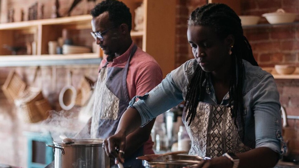 Un couple cuisine dans un espace qui semble assez grand. Ils s'affairent devant des casseroles et une plaque de cuisson.