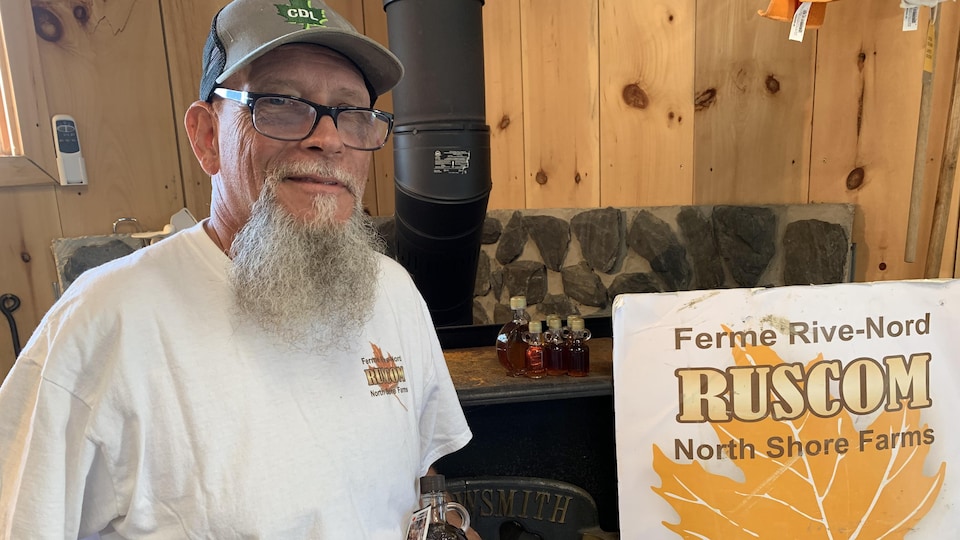 Un homme avec une barbe, portant une casquette, tient dans ses mains une bouteille de sirop d'érable et d'autres produits d'érable