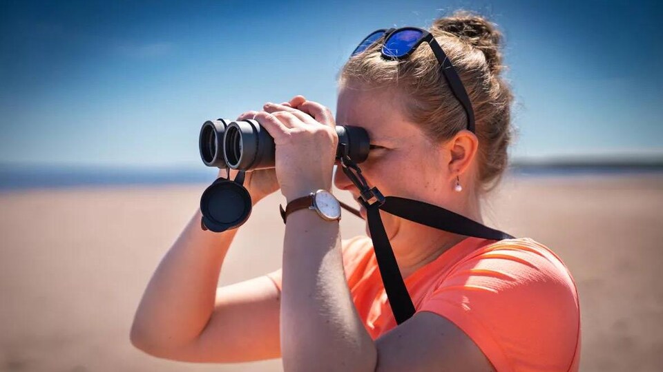 Sarah Hirtle vue de profil sur une plage, regardant au loin dans des jumelles.