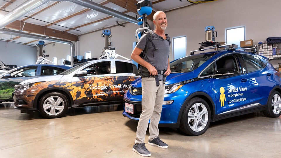 Un homme porte un sac à dos contenant une caméra, et se tient debout dans un garage rempli de voitures à l'effigie de Google Street View. 