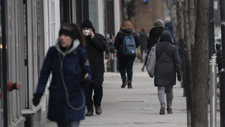 Plusieurs personnes, dont une porte un masque, marchent sur un trottoir.