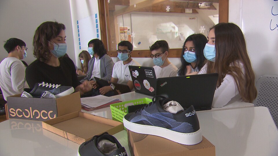 Des chaussures sont posées sur une table. Des élèves sont assis tout près, portant des masques, certains avec des ordinateurs portables.