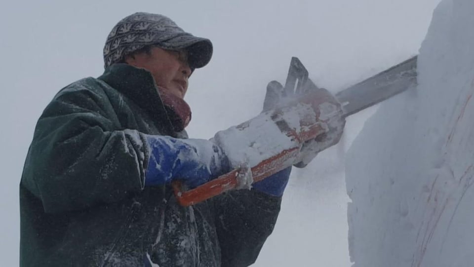 Gros plan sur un homme de profil habillé très chaudement et qui manie une scie pour façonner une sculpture de neige. Son manteau est couvert de neige et de la neige poudreuse revole dans son visage.
