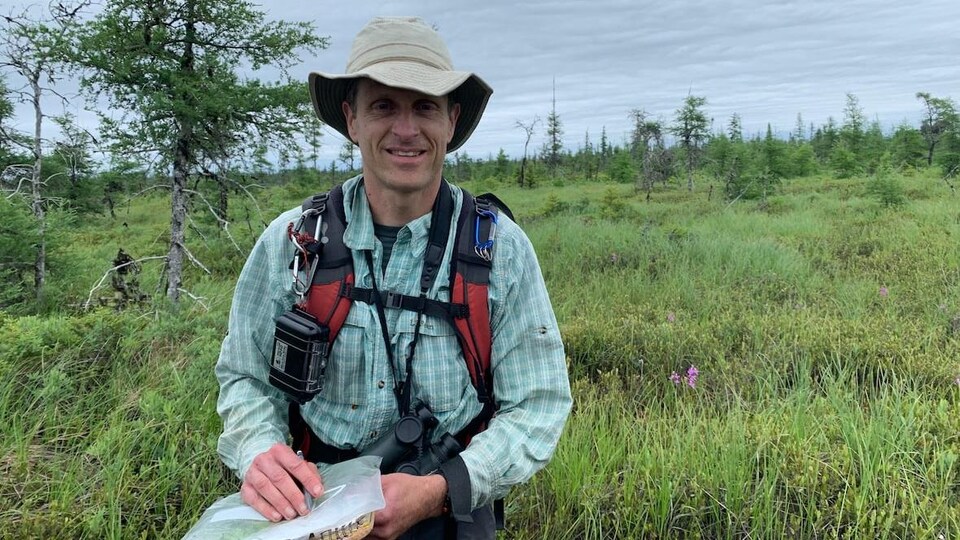 Sean Blaney vêtu de son équipement de recherche tient des outils dans ses mains. Il est photographié dans les herbes hautes de la tourbière d'Alward Brook.