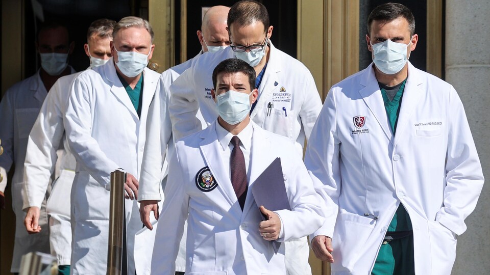 Une équipe de médecins en sarrau s'avance vers les journalistes.