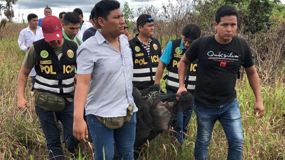 Des policiers habillés en civil, mais certains avec une veste sur laquelle il est inscrit POLICIA, transportent le corps d'un homme décédé enveloppé dans une toile. Ils sont dans un champs avec des herbes hautes.