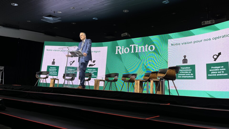 Sébastien Ross devant un podium sur une scène aménagée avec des chaises pour une discussion de groupe en public.