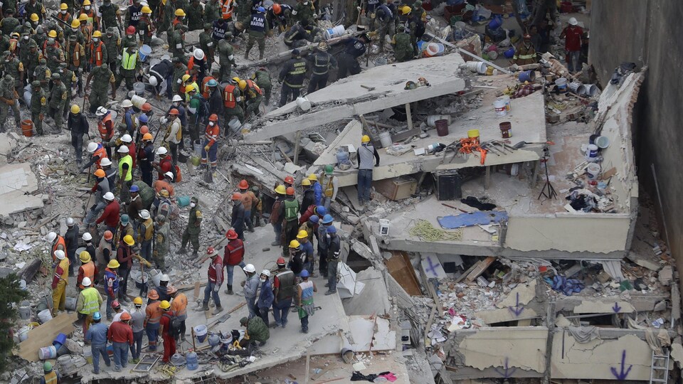 Des dizaines de travailleurs portant des casques de protection sont au milieu de ruines.