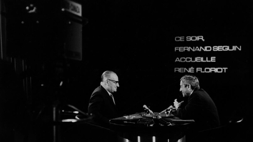 Dans un studio de télévision, René Floriot s'entretient avec l'animateur Fernand Seguin. Deux microphones sont posés sur le pupitre devant eux.