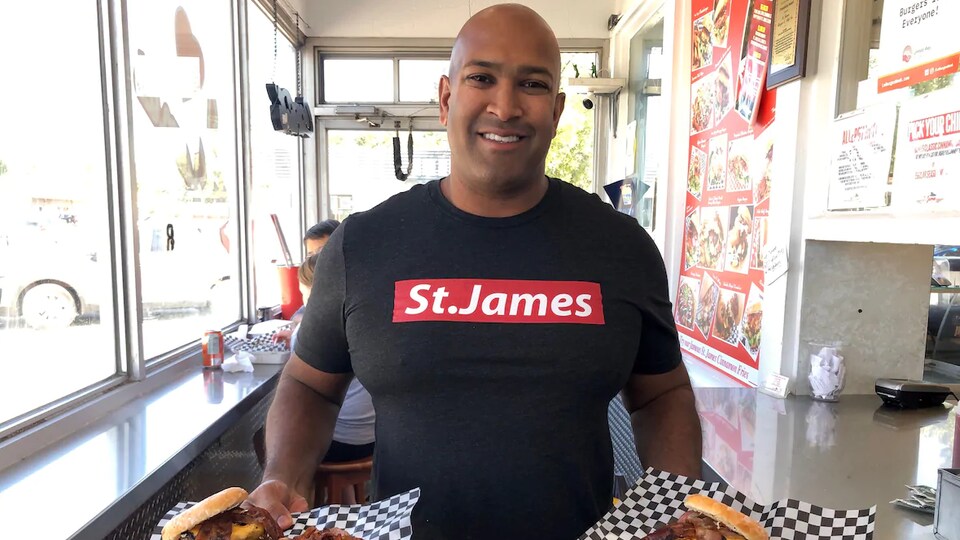 Un homme portant un chandail noir où est inscrit les mots St.James avec deux assiettes contenant des hamburgers et des frites dans ses mains.