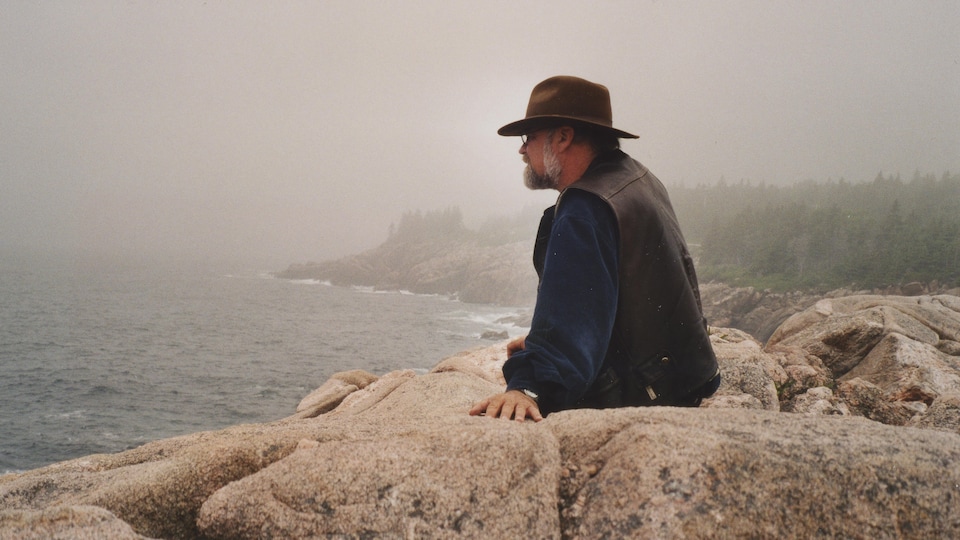Serge Bouchard assis sur des rochers, regardant le fleuve en Nouvelle-Écosse, près de Cabot Trail.