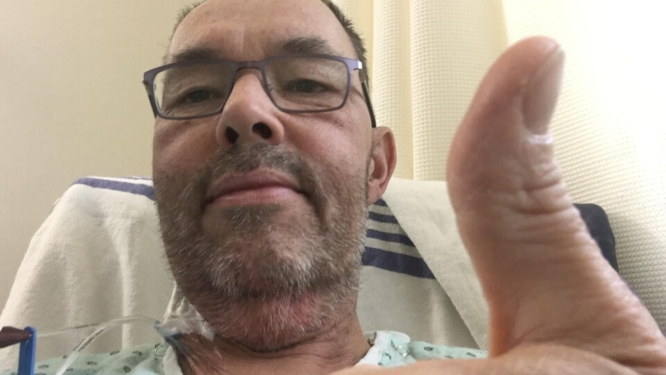 Serge Dion, dans un lit d'hôpital, lève un pouce en l'air.