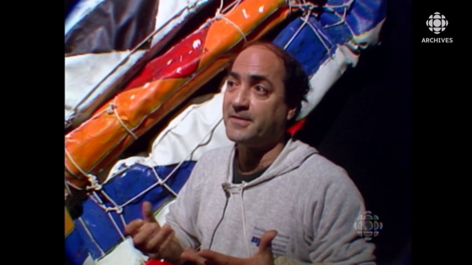 Le peintre Serge Lemoyne qui parle devant des toiles peintes enroulées de ficelles. 