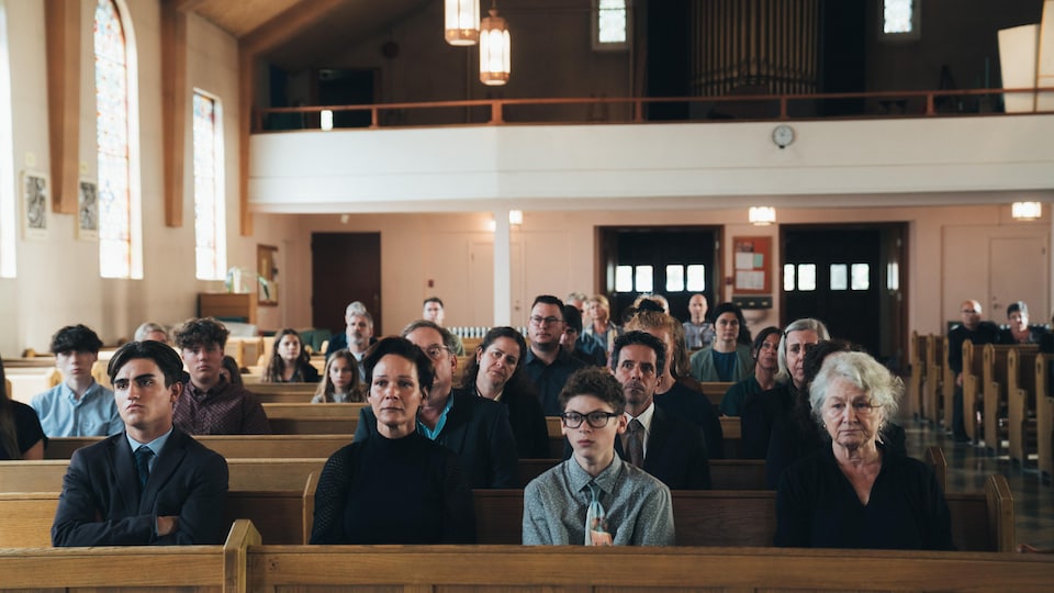 Des gens vêtus d'habits funéraires sont assis dans des bancs d'église. 