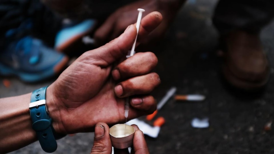 Zoom sur des mains sales qui utilisent une seringue pour extraire de la drogue d'un petit contenant. En arrière-plan des seringues traînent sur le sol.